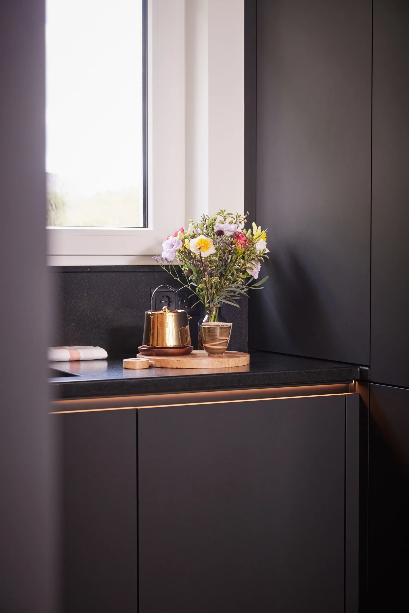 Cuisine moderne avec plan de travail en marbre noir, bouquet de fleurs et pot en cuivre près de la fenêtre. Ambiance élégante et lumineuse.