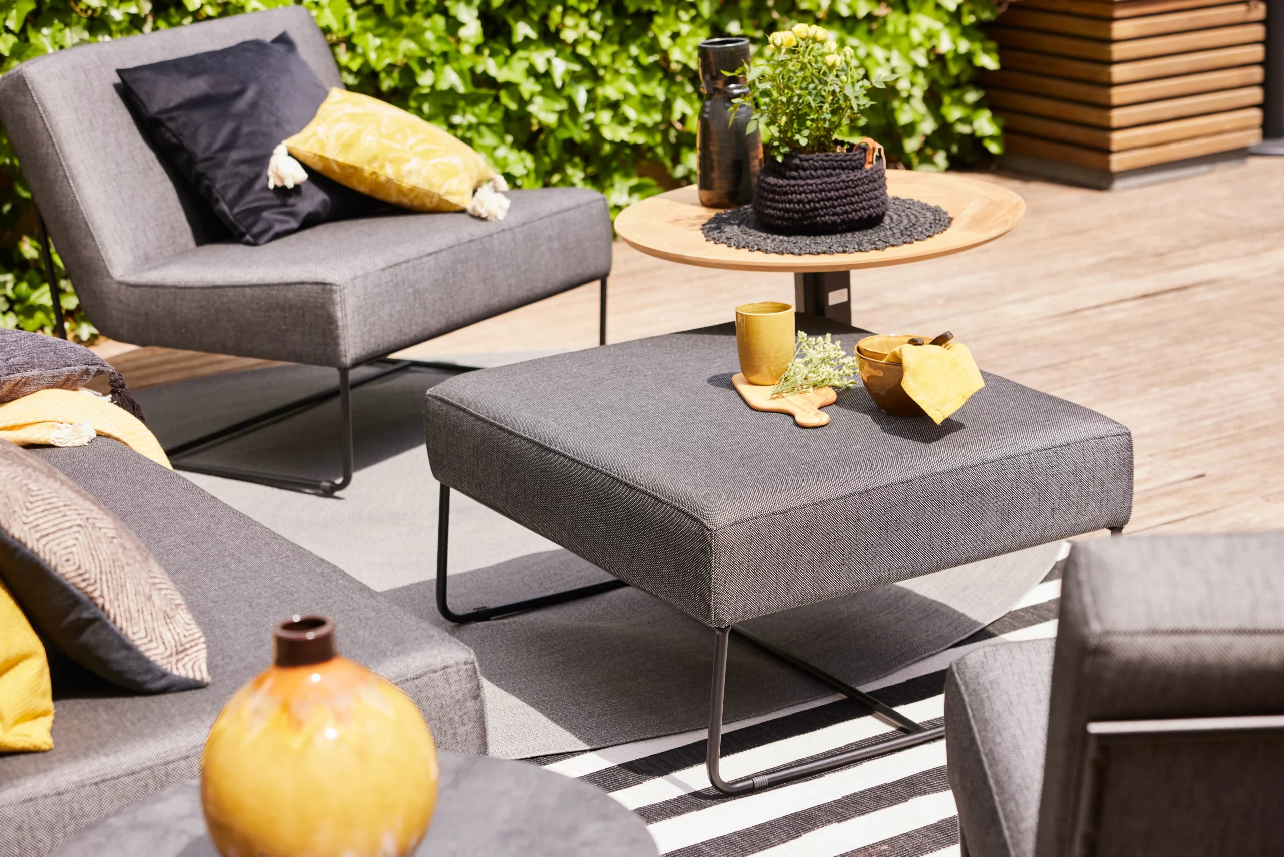 Mobilier de jardin moderne avec fauteuils gris et table en bois, décoré de coussins colorés et plantes vertes sur fond de terrasse ensoleillée.