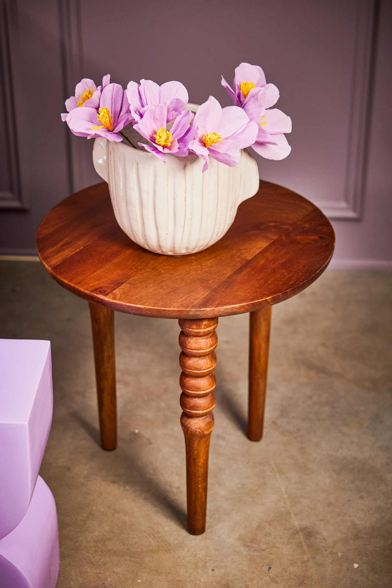 Vase en céramique blanche avec fleurs violettes sur une table en bois, contre un mur lilas.