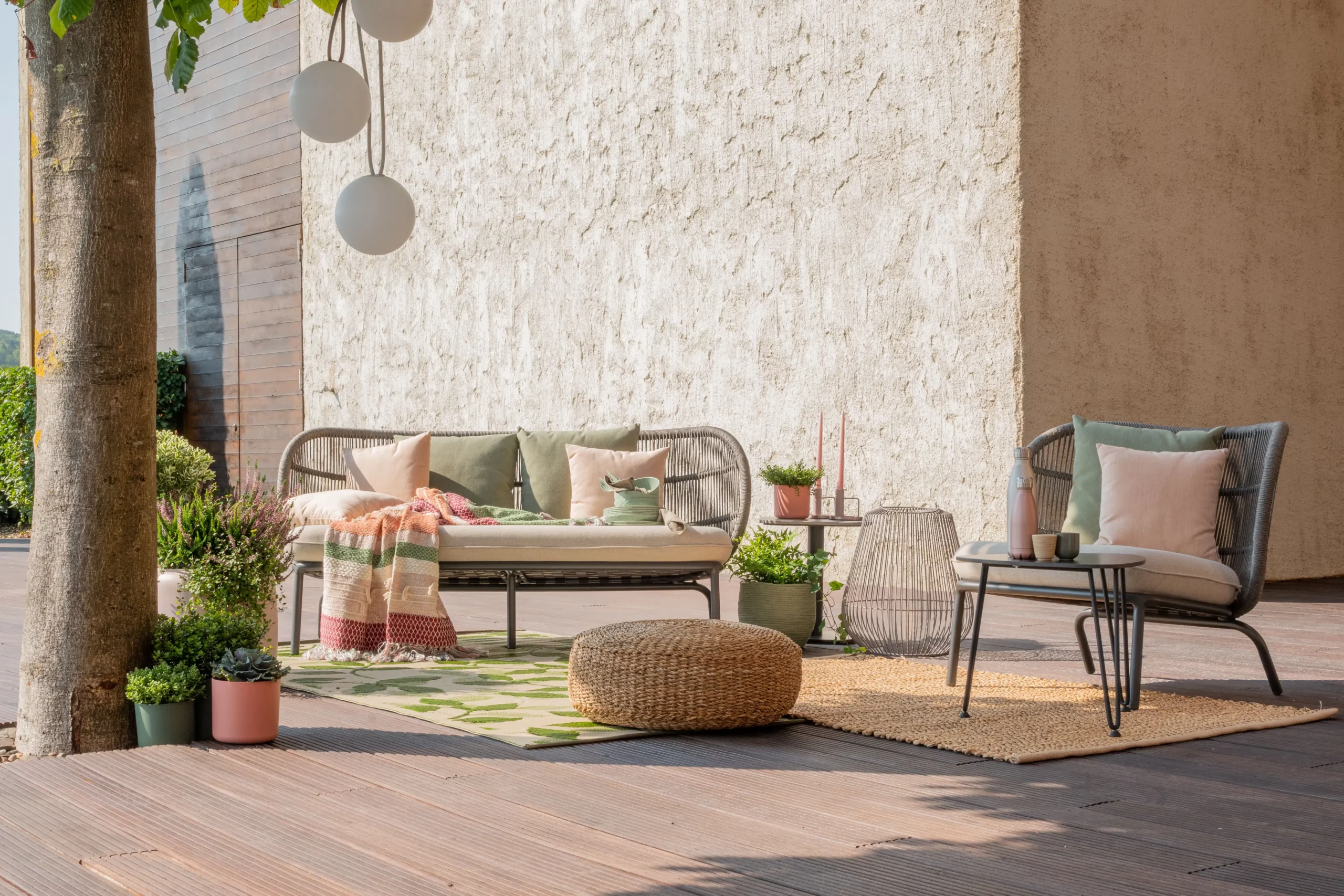 Aménagement terrasse chic : mobilier en rotin, tapis végétal, pouf tressé et plantes, créant un espace extérieur chaleureux et design sous un arbre ombragé.